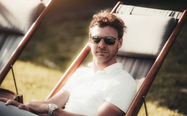 Kıvırcık kahverengi saçlı ve beyaz polo gömlekli beyaz bir adam güneşli bir akşamda açık havada bir iskemlede oturuyor. Güneş gözlüğü ve kol saati takıyor. Solda bir kopyalama alanı