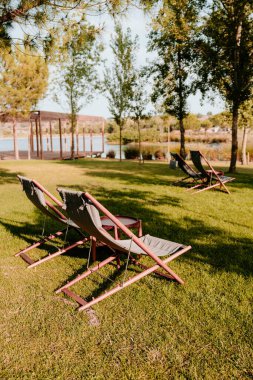 Ağaçlar ve gölle çevrili yeşil bir çimenlikteki ahşap şezlonga sahip huzurlu bir açık hava manzarası. Bölge yumuşak güneş ışığıyla yıkanmış, sakin ortamın altını çiziyor.