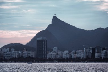 Rio de Janeiro 'nun kıyı şeridinin panoramik bir görüntüsünde Corcovado Dağı' ndaki ikonik İsa heykeli, gökyüzü alacakaranlıkta pastel renkli bir gökyüzünün altında ufuk çizgisi ve binalar var.