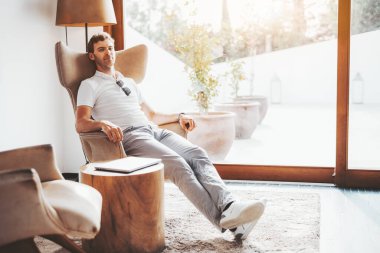 Beyaz polo gömlekli ve gri pantolonlu beyaz bir adam geniş bir pencerenin yanındaki modern bir koltukta dinleniyor. Güneş ışığı içeri akıyor, sakin ve minimalist kapalı mekan ayarlarını vurguluyor.