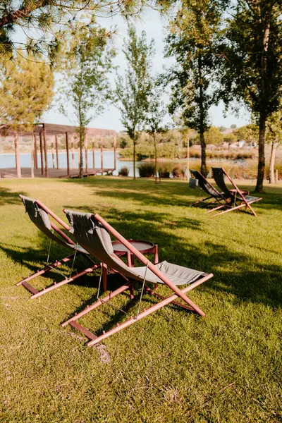 Ağaçlar ve gölle çevrili yeşil bir çimenlikteki ahşap şezlonga sahip huzurlu bir açık hava manzarası. Bölge yumuşak güneş ışığıyla yıkanmış, sakin ortamın altını çiziyor.