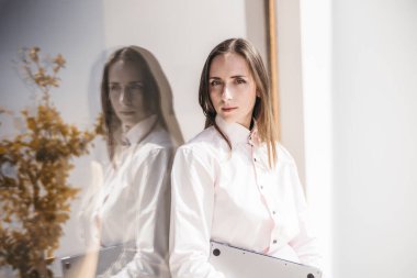 Beyaz bluzlu ve pembe etekli bir kadının portresi dizüstü bilgisayar tutuyor, cam bir pencerenin yanında duruyor ve görüntüsünü yansıtıyor. Arka planda saksı bitkileri ve beyaz duvarlar