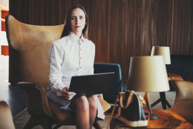 Beyaz gömlekli ve eflatun etekli İspanyol iş kadını modern bir koltukta dizüstü bilgisayarla oturuyor. Arkaplanda, profesyonel ve şık bir atmosfer yaratan ahşap bir duvar ve zarif bir dekor yer alıyor.