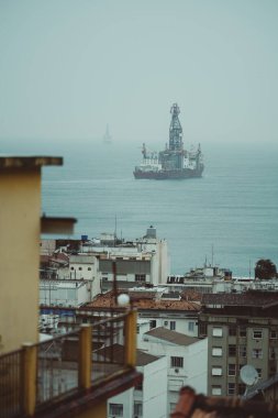 Sisli ufukta görünen iki kıyı sondajı ile okyanusun insansız hava aracından düşey bir telsiz görüntüsü, ön planda yerleşim binaları olan bir şehir manzarasından görülüyor.