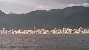 Rio 'nun Ipanema plajının panoramik manzarası ve sudan ele geçirilen yüksek binaları. Sahne, hareketli şehir manzarası ile arkadaki dingin dağlar arasındaki zıtlığı gösteriyor.