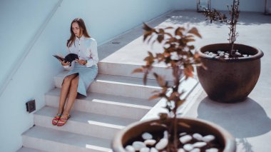 Beyaz gömlekli ve açık etekli bir kadın açık hava basamaklarında oturup kitap okuyor. Minimalist ortamı taşlı saksı bitkileri süslüyor. Sahne yumuşak, doğal bir ışıkla çekiliyor.
