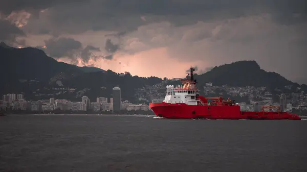 stock image A large red offshore supply vessel navigates through the waters near Rio de Janeiro, with the city's skyline and mountainous landscape visible in the background during a dramatic sunset