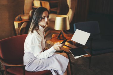 Beyaz gömlekli ve lavantalı etekli bir iş kadını modern bir salonda elinde akıllı bir telefonla oturuyor. Bir dizüstü bilgisayar, defter ve lamba yakınlardaki ahşap bir masada.