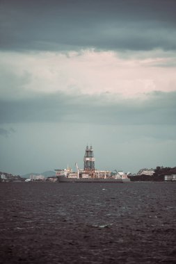 A deepwater drillship stationed offshore under overcast skies. The vessel features a prominent drilling tower and industrial structures, set against a distant coastal cityscape clipart