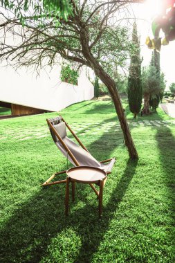 Ahşap bir iskemle ve yemyeşil bir çimenlikte küçük yuvarlak bir masa içeren sakin, dikey bir açık hava sahnesi. Sandalye yakındaki bir ağaçla gölgelenmiş, benekli güneş ışığı huzurlu bir ortam yaratıyor.