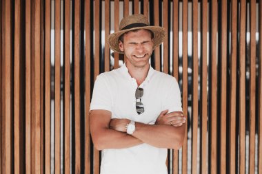 Hasır şapkalı ve beyaz polo tişörtlü gülen bir adam, kolları çaprazlanmış, tahta bir duvarın önünde duruyor. Parlak, doğal ışık rahatlatıcı, neşeli atmosferi güçlendirir.