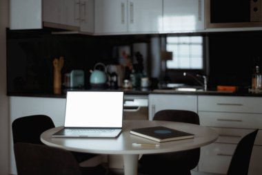 Modern bir mutfaktaki yuvarlak bir masada boş beyaz ekranlı, dizüstü bilgisayarın gösterişsiz bir görüntüsü. Yanında da arka planda defter, kalem ve minimalist dekor unsurları var. Şablon kullanımı için ideal