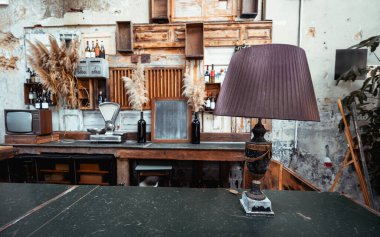 Ön planda kıvrımlı bir gölgesi olan klasik bir masa lambasının olduğu şık bir bar tezgahı. Arka planda ahşap rafları, şarap rafı ve vazolarda otlar bulunan rustik dekorlar sergileniyor.