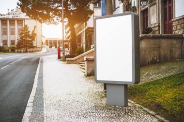 Gün batımında Avrupa 'da bir şehrin kaldırımındaki kaldırımda boş bir reklam panosu duruyor. Arka planda binalar ve ağaçlar görünür.