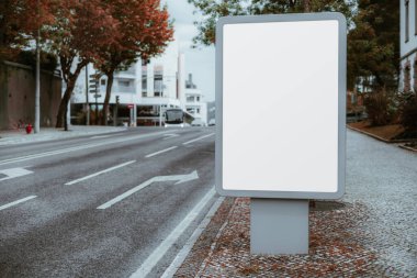 Boş beyaz ekranlı boş dikey bir reklam panosu kaldırımda duruyor, trafiği olmayan sakin bir şehir yolunun yanında, ağaçlar ve modern binalarla çevrili.