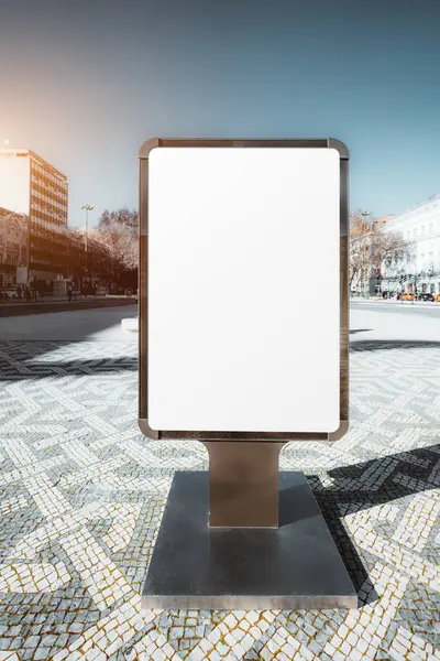 stock image Empty vertical billboard with a blank white surface in a modern urban setting, situated on a patterned stone pavement. The surrounding area includes buildings and trees under a clear sky