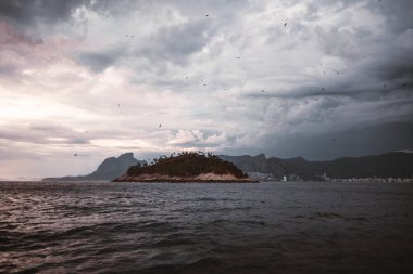 Brezilya 'nın Rio de Janeiro kıyısında Atlantik Okyanusu ile çevrili küçük yemyeşil bir ada. Tepemizde dramatik bulutlar belirdi uzak şehir silüeti ve dağlarla