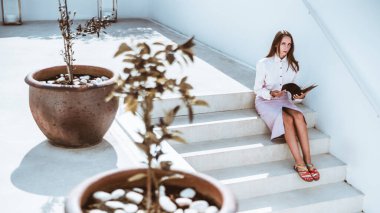 Açık pembe bluz ve mor etekli genç bir kadın açık bir defter tutan beton basamaklarda oturuyor. Sahne, merdivenin her iki tarafında da büyük saksı bitkileriyle aydınlık.