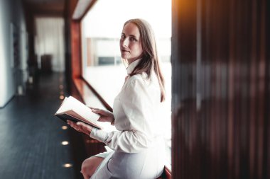 Uzun düz saçlı, beyaz bluz ve açık etek giyen bir kadın, modern bir iç mekanda geniş bir pencerenin yanında oturuyor. Kameraya bakarken açık bir kitap tutuyor.