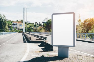 Boş bir reklam panosu modeli köprünün yakınındaki bir yol kenarında belirgin bir şekilde duruyor. Yol boş, güvenlik rayları, ağaçlar ve manzaralı arka planda uzak evler var.