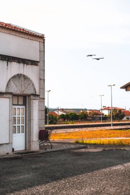 Güneşli bir şehir manzarası, kapalı kapısı, park etmiş alışveriş arabası ve havada uçan iki kuşu olan eski beyaz bir binanın bir kısmını gösteriyor. Arka planda demiryolu rayları ve evler görünüyor.