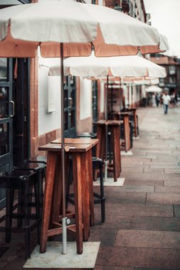 Şehir caddesinde ahşap masaları, bar tabureleri ve beyaz şemsiyeleri olan açık hava kafesi. Sığ alan derinliği uzak sokağı vurguluyor. Öndeki masaya seçici odaklanma