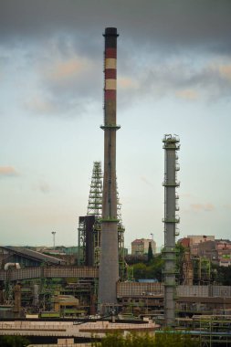 Şehir binalarıyla çevrili iki uzun baca ve karmaşık rafineri yapısına sahip bir sanayi manzarasının telefoto görüntüsü. Bulutlu gökyüzü karamsar, atmosferik bir dokunuş ekliyor.