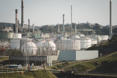 Geniş bir endüstriyel petrol rafinerisi geniş küresel ve silindirik depolama tankları, çoklu boru hatları ve temiz bir gökyüzünün altındaki yeşil tepelerle çevrili uzun bacalar.