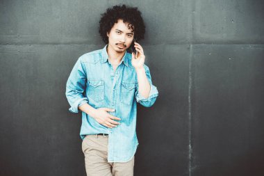 Sıradan mavi kot gömlekli kıvırcık saçlı genç adam, kulağına akıllı telefon dayamış, karanlık bir duvarın karşısında duruyor. Bir sohbete odaklanmış ve meşgul görünüyor.
