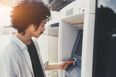 Kıvırcık saçlı genç Asyalı bir adam ATM 'ye açık tişört ve siyah tişört giyerek bir banka kartı sokuyor. Dış mekan, işletim sürecine odaklanarak güneş ışığıyla aydınlatılıyor.