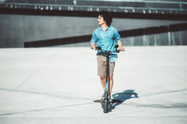 Kıvırcık saçlı genç bir adam elektrikli scooter 'ın üzerinde açık mavi kot gömlek ve haki rengi şort giyiyor. Minimalist, kentsel bir geçmişe karşı yan gözle bakıyor.