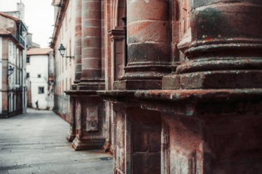 Tarihi bir binanın ön cephesindeki yıpranmış taş sütunların yakın görüntüsü, detaylı doku ve mimari öğeler sergileniyor. Arka planda sessiz dar bir cadde soluyor