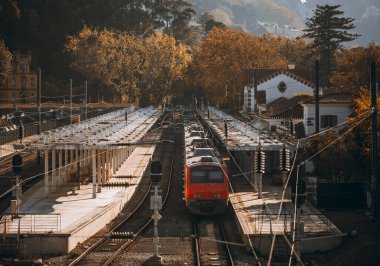 Kırmızı bir tren, bereketli sonbahar yeşillikleri, beyaz geleneksel Avrupa mimarisi ve yumuşak, altın renkli güneş ışığıyla çevrili sessiz ve manzaralı bir tren istasyonuna varır.