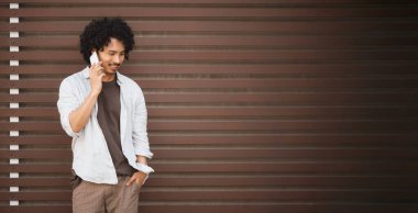 Beyaz gömlek ve kahverengi şort giyen kıvırcık saçlı genç bir koridor adamı modern kahverengi bir duvarın önünde duruyor ve rahat bir ifadeyle akıllı telefonuyla konuşuyor. Boşluğu sağda kopyala