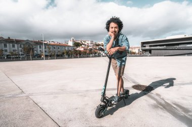 Günlük kıyafetli kıvırcık saçlı bir adam büyük, boş bir şehir meydanında elektrikli bir scooter 'ın yanında düşünceli bir şekilde duruyor. Bir eli çenesinde, parçalı bulutlu bir gökyüzünün altında scooter 'a yaslanıyor.