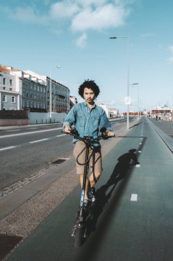 Modern Avrupa şehrinde boş bir bisiklet yolunda elektrikli scooter kullanan kıvırcık saçlı genç bir adam. Kot gömlek ve ekose şort giyiyor.