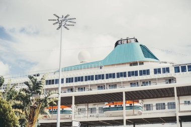 Limana yanaşmış büyük, modern bir yolcu gemisinin yakın çekimi. Pencereleri, filikaları, uzun bir lamba direği ve bulutlu bir gökyüzüne karşı kurulmuş tropikal yaprakları olan güverteler görülebilir durumda.