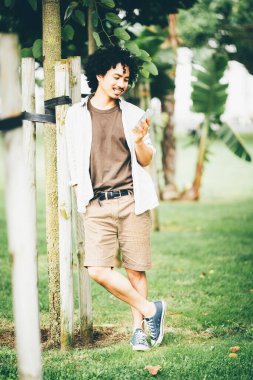 Kıvırcık saçlı, günlük kıyafetler giyen, parkta dikilen genç bir adam. Ağaca yaslanıyor, elinde gülümsemeli bir akıllı telefon, şort, tişört ve spor ayakkabı tutuyor.