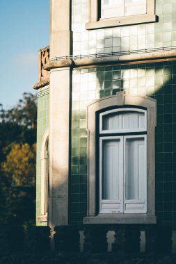 Yeşil seramik fayanslara sahip tarihi bir binanın cephesi, karmaşık mimari ayrıntılar, ve güneş ışığını yansıtan beyaz çerçeveli pencereler; süslü korkulukları olan bir balkon