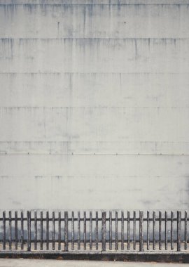 Minimalist şehir kompozisyonu, dokusu gri beton bir duvar görünür hava lekeleri ve ön planda seçici bir şekilde odaklanmış siyah metal bir çit.