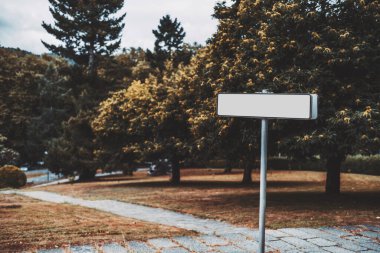 Metal bir direğe monte edilmiş boş beyaz dikdörtgen bir tabela manzaralı bir parkta duruyor, ağaçlarla çevrili, taş bir patika, ve sonbahar yeşillikleri olan çimenlikli bir alan.