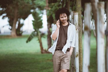 Akıllı bir telefonla konuşan kıvırcık saçlı genç bir adamın, yeşil ve ağaçlarla çevrili bir parktaki ahşap bir direğe yaslanırken çekilmiş bir fotoğrafı.