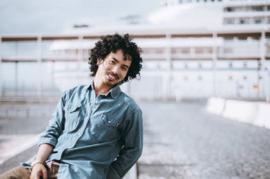 Kıvırcık siyah saçlı ve bıyıklı genç bir adam sıradan gülümsüyor, kot gömlek ve kahverengi pantolon giyiyor, endüstriyel veya kentsel bir ortamda açık havada poz veriyor.