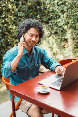 Kot gömlek giyen kıvırcık saçlı melez bir adamın dikey görüntüsü kırmızı bir masada oturup dizüstü bilgisayarda çalışıyor, akıllı bir telefon tutuyor ve bir fincan espressonun tadını çıkarıyor.