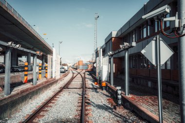 Güvenlik kameraları, bariyerler ve bitişik modern ofis binası olan bir sanayi bölgesinde güvenli bir demiryolu hattı. Üst geçit, park edilmiş arabalar ve arkada nakliye konteynırları.