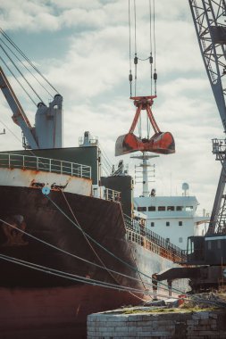 Limana yanaşan endüstriyel kargo gemisinin dikey telefoto görüntüsü. Büyük paslı vinç gövdeyi boşaltıyor. Ağır donanım, deniz lojistiği ve küresel nakliye endüstrisi konsepti