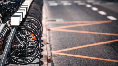 Park halindeki kiralık bisikletlerin sırası turuncu işaretli bir yolun yakınındaki bir rıhtım istasyonunda. Ön tekerleklerin yakın çekimi ve keskin odaklı teller, arka plan bulanık. Boşluğu sağda kopyala