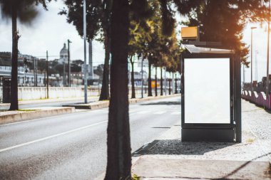 Boş bir şehir sokağındaki otobüs durağında reklam panosu modeli boş. Ağaçlar, yol işaretleri ve tarihi mimarisi yumuşak odaklı bir şehir manzarası solda bir kopya alanı.