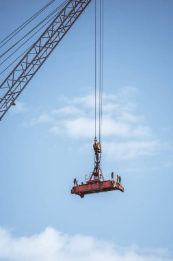 Ağır bir endüstriyel vinç, dağınık bulutlu parlak mavi gökyüzüne karşı birden fazla mandalı ve mekanizması olan kırmızı bir çelik yayıcı çubuğu kaldırıyor. Kablolar ve makara sistemi görünür