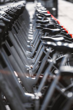 Bir sıra modern kiralık elektrikli bisiklet düzgünce bir bisiklet paylaşım istasyonuna kenetlendi. Görüntü siyah eyerler ve metalik çerçeveler üzerinde yoğunlaşarak, yinelenen bir kentsel şablon oluşturur; telefoto çekimi